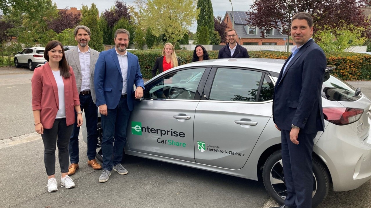 Seven people standing around a silver Enterprise Car Share vehicle and looking at the camera