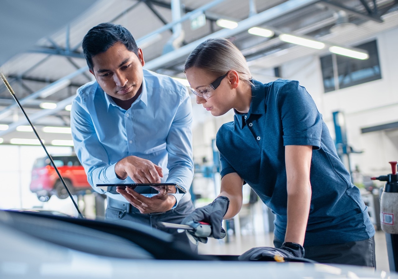 Instructor with a Tablet Computer is Giving a Task for a Future Mechanic. Female Student Inspects the Car Engine. Assistant is Checking the Cause of a Breakdown in the Vehicle in a Car Service.